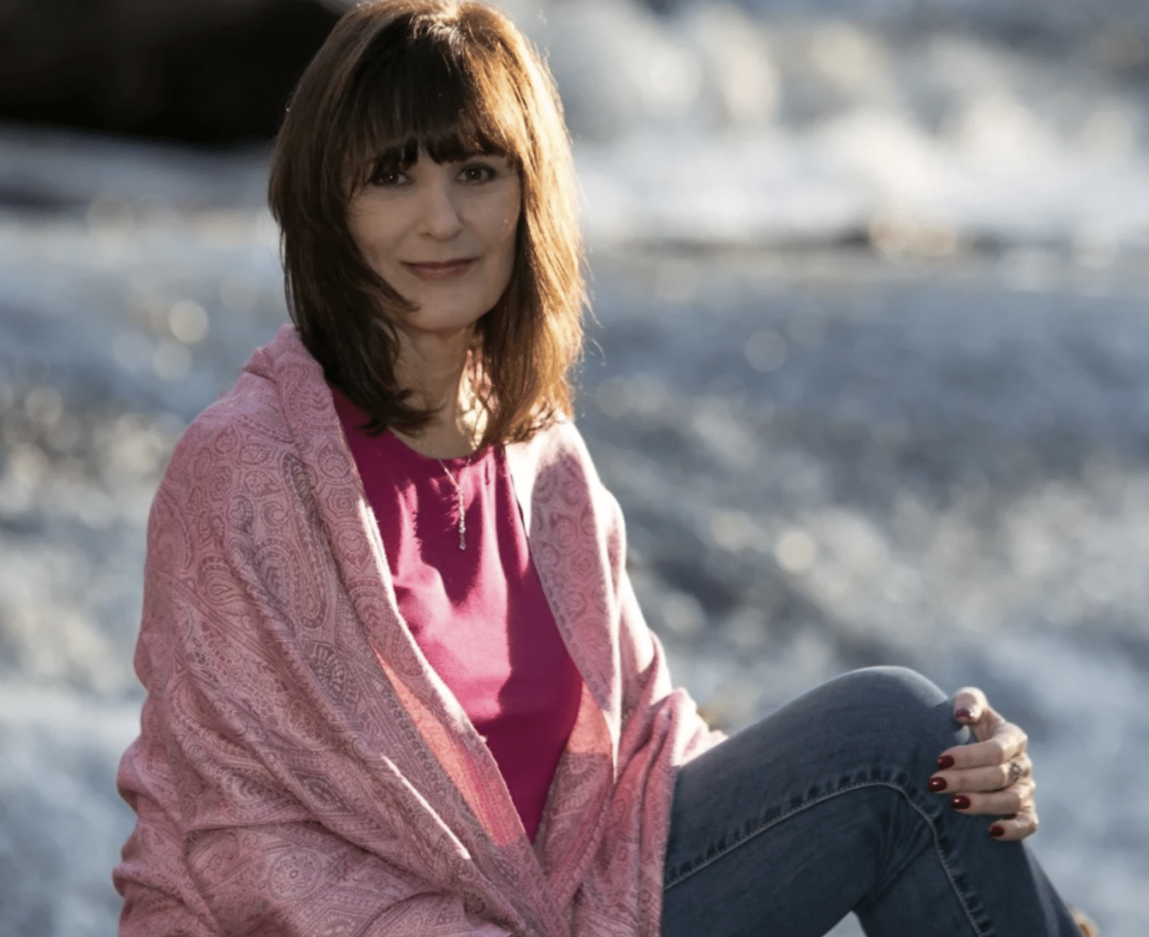 Author Tammy Billups sits on the beach with the sun shining off her hair and wearing a soft pink shawl
