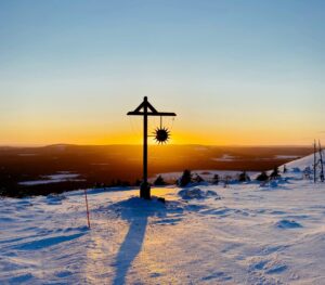 The image captures a serene winter scene at sunset, atop a snow-covered hill. In the foreground, there's a wooden street light adorned with a sun-shaped ornament. The sun is positioned directly behind the ornament, creating a silhouette effect and a burst of light.