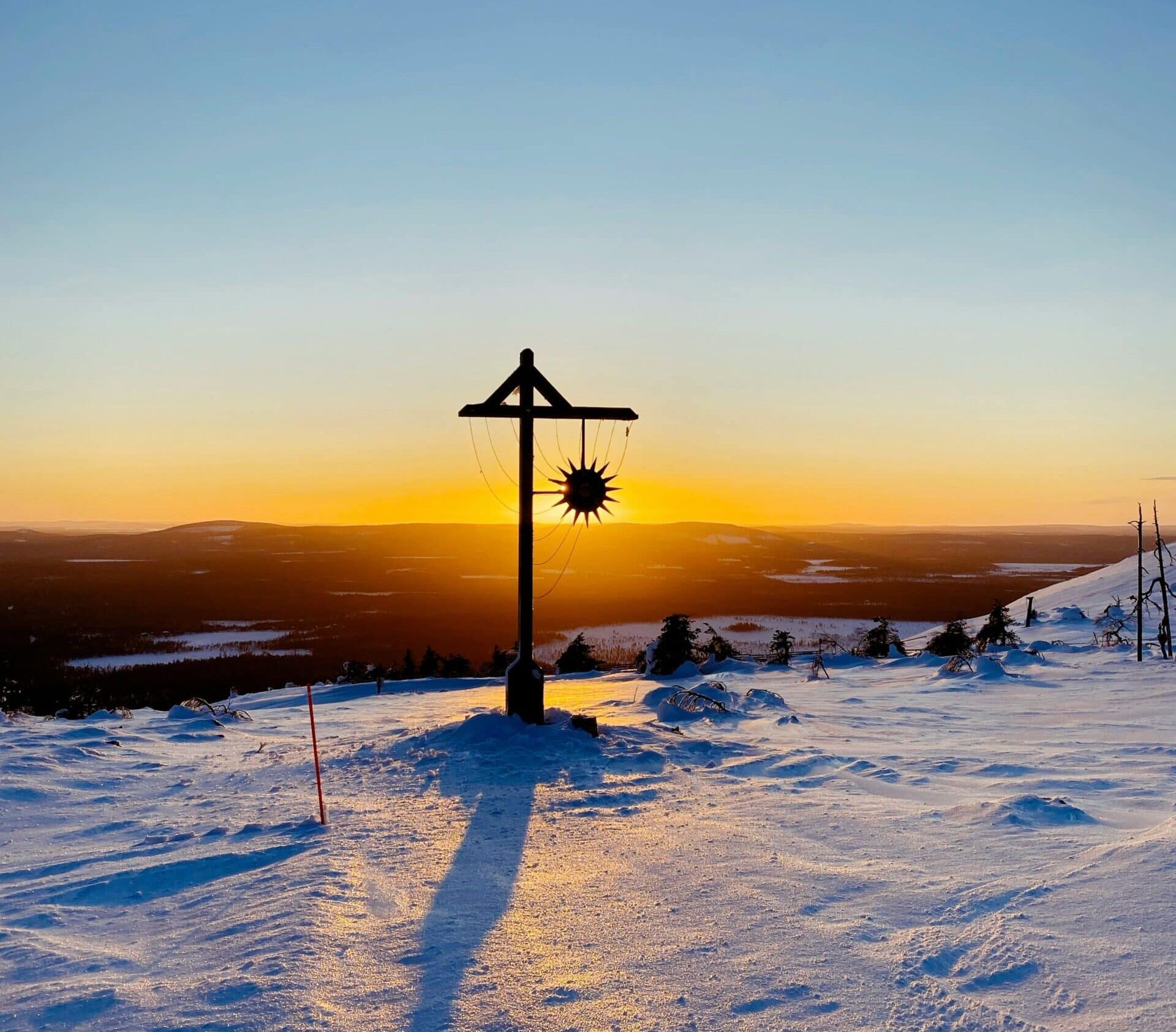 The image captures a serene winter scene at sunset, atop a snow-covered hill. In the foreground, there's a wooden street light adorned with a sun-shaped ornament. The sun is positioned directly behind the ornament, creating a silhouette effect and a burst of light.